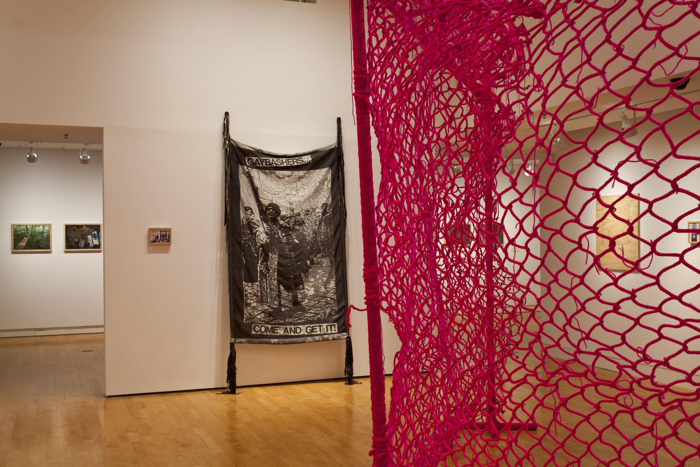 Installation shot: Foreground: We Couldn’t Get In. We Couldn’t Get Out. (2006-2007); Crank-knit yarn, hand-woven wire, steel poles. Midground: Gay Bashers Come And Get It (2011); Jacquard-woven cotton and lurex, hand-dyed fabric, crank-knit yarn, thread. Graphic appropriated from a poster designed by Matt Height, silkscreened at Queeruption (1999) at DUMBA, Brooklyn, and used as an album insert for queercore band Limp Wrist. Both by L.J. Roberts and courtesy of the artist.