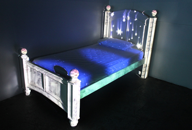 A white bedframe with purple bed, lights being projected onto the headboard